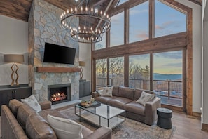 A spacious living room with a stone fireplace, leather sofas, a glass coffee table, and large windows offering a scenic view.