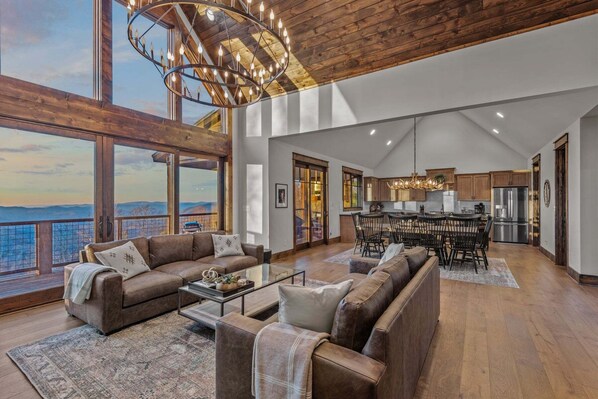 A spacious living area with a large window, a chandelier, and a dining area with wooden cabinets.
