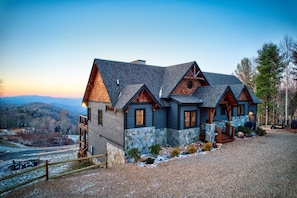 A large, multi-story house with a stone foundation and wooden exterior, set in a mountainous area with a gravel driveway and a wooden fence.