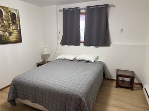 A bedroom with a bed, two windows with curtains, a bedside lamp, a small wooden nightstand, and a painting on the wall.