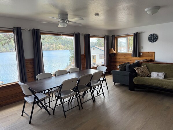A room with a dining table, chairs, a sofa, a ceiling fan, and large windows with curtains.