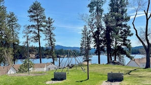 A grassy area with swings, a wooden structure, and a lake with trees in the background.