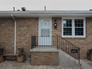 A brick house with a white door, a small porch with a bench, a window, and a mailbox.