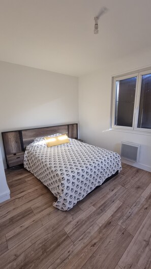 Une chambre avec un plancher en bois, un lit recouvert d’une couvre-lit à motifs, une table de chevet, une fenêtre avec des stores et un luminaire au plafond.