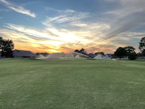 A golf course with a sunset in the background, houses in the distance, and a sprinkler system in the foreground.