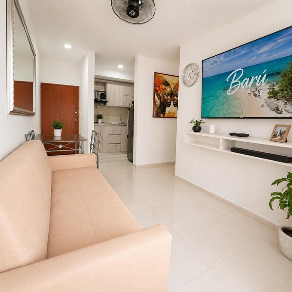 Un salon avec un canapé beige, un téléviseur diffusant une scène de plage, une cuisine en arrière-plan et un coin repas avec une table et des chaises.