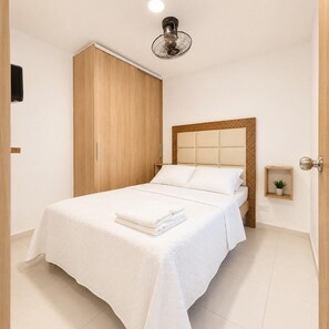 Un lit bien fait, recouvert d’une courtepointe blanche et agrémenté d’oreillers, une armoire en bois, une table de chevet avec une plante et un ventilateur de plafond.