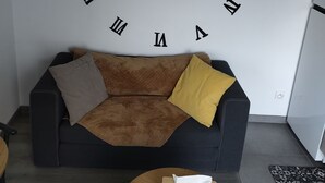 A living room with a large wall clock, a brown sofa with cushions, a round wooden coffee table with a tissue box, and a patterned rug. - Charming full T1 (Zillisheim)