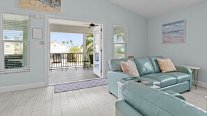A living room with a light blue sofa, a small table, and a balcony with a view of houses and palm trees. - Dolphin Run | Heated Pool, 3-Min Walk to Beach! (Fort Myers Beach)