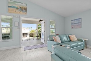 A living room with a light blue sofa, a small table, and a balcony with a view of houses and palm trees. - Dolphin Run | Heated Pool, 3-Min Walk to Beach! (Fort Myers Beach)