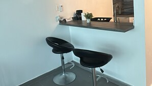 Et moderne køkken med indbygget ovn, et ur og en velkomstskilt. Der er to barstole og en lille urtepotte på bordpladen.