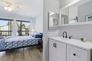 Ein Schlafzimmer mit Bett, Deckenventilator, großem Fenster mit Vorhängen, Bad mit Waschbecken, Spiegel und Schränken.