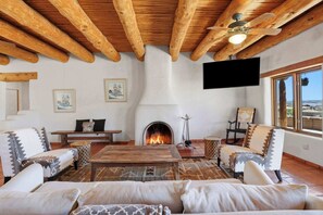Ein gemütliches Wohnzimmer mit Kamin, Holzbalkendecke, Fernseher und einem großen Fenster.