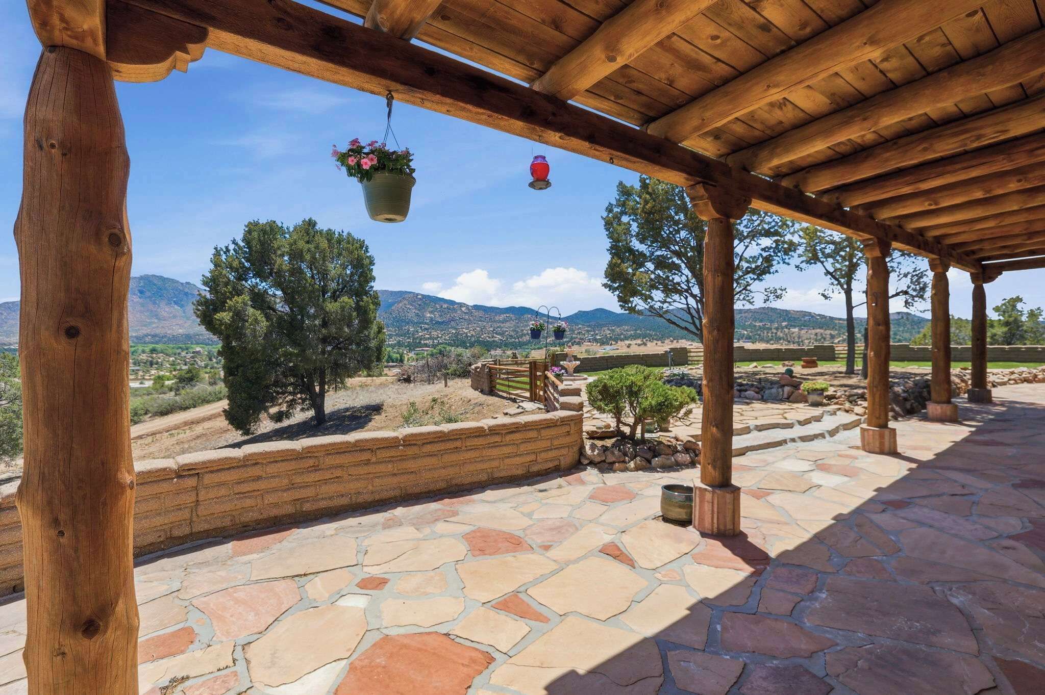 A covered patio with stone flooring, wooden pillars, hanging flower pots, and a view of distant mountains.