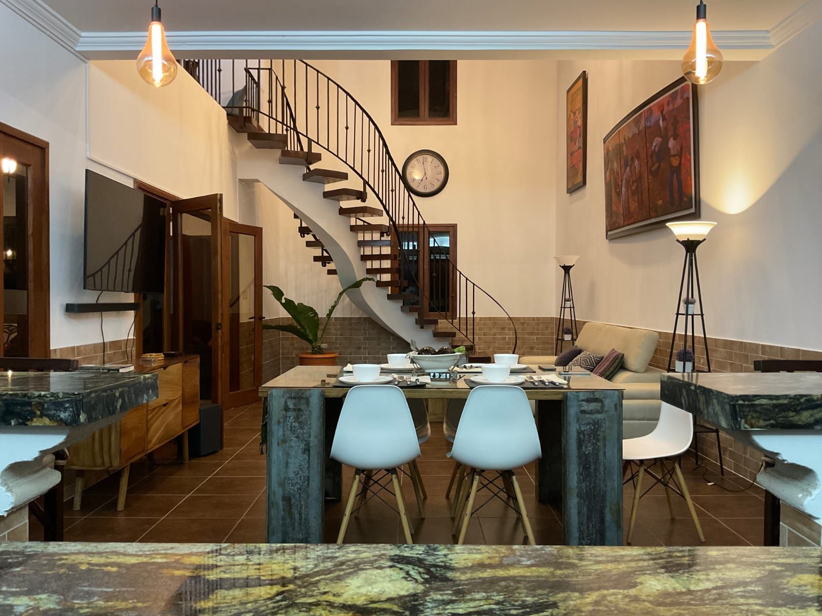 Une cuisine moderne avec un îlot central, un coin repas avec des chaises, un escalier et une horloge au mur.