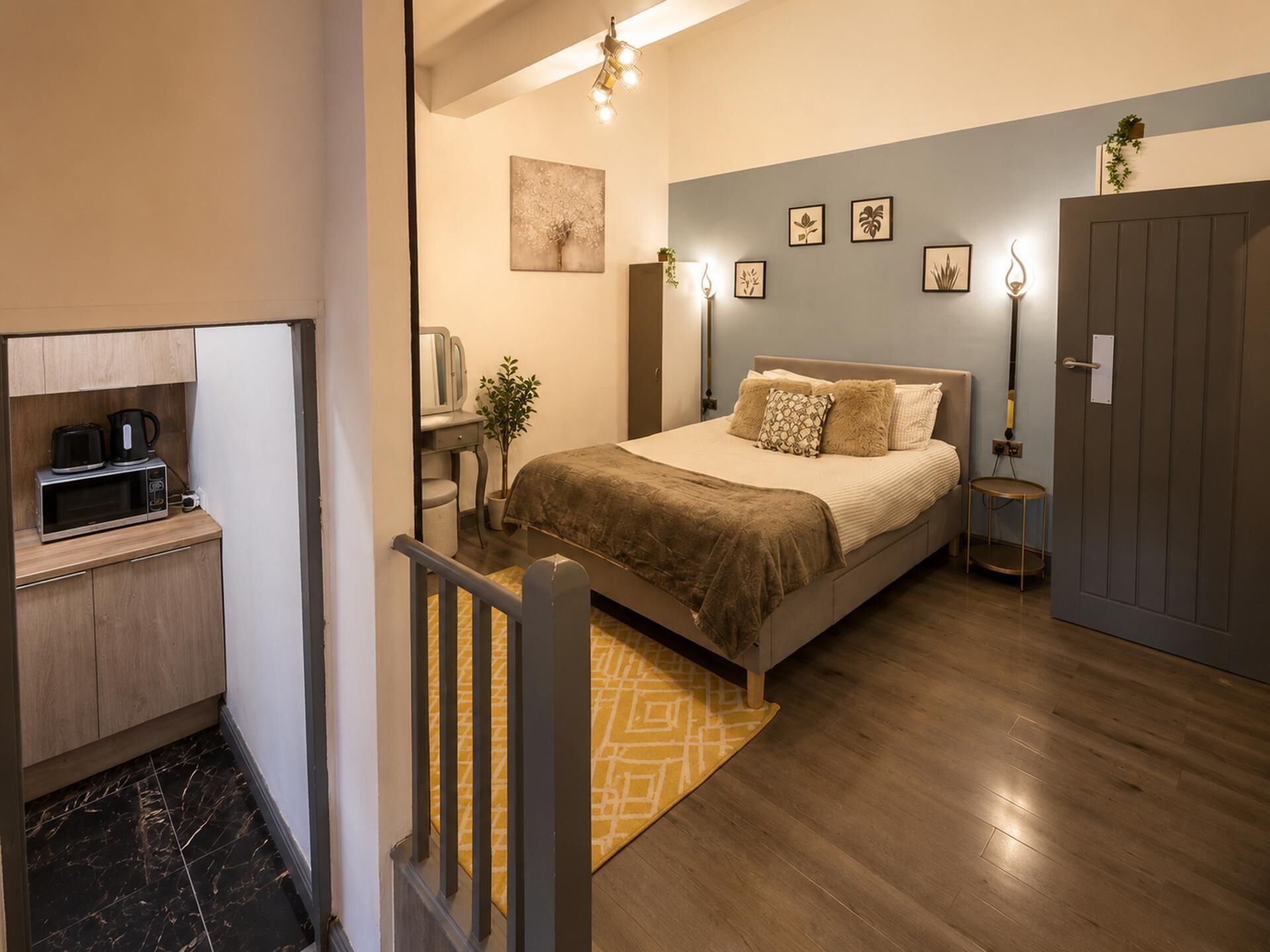 Une chambre à coucher avec un lit, des tables de chevet, un bureau avec une chaise, une petite plante et une porte.