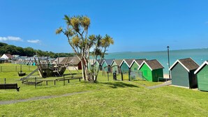 A row of green beach huts with a clear view of the sea and a tree in the foreground. - Stylish Coastal Cottage in Gurnard, Dog Friendly Garden (Cowes)
