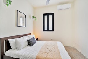 Een slaapkamer met een bed, een nachtkastje, een wandairconditioner, een raam en een lijst met een afbeelding.
