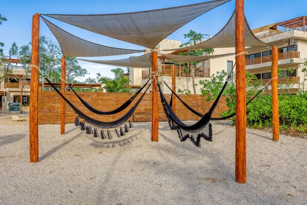 A hammock under a shaded area with a sandy ground, wooden posts, and a building in the background. - Boho Apartment with Pool & Cenotes  (Tulum)