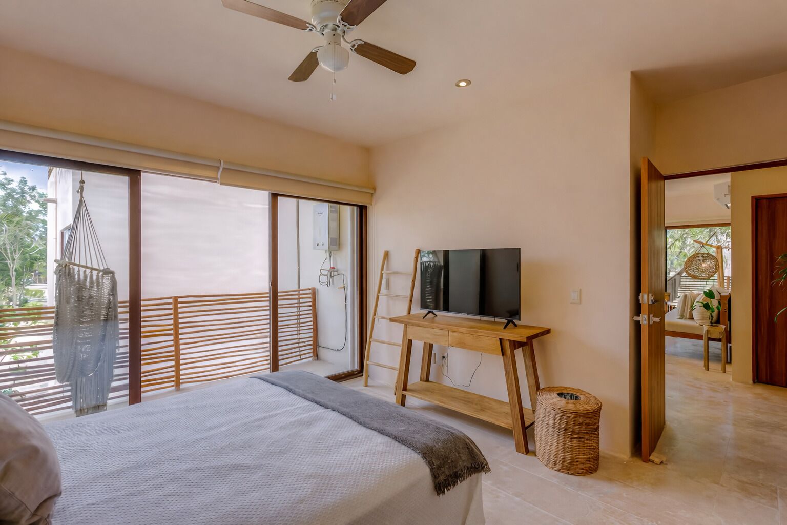 Une chambre à coucher avec un lit, un téléviseur posé sur un meuble en bois, un ventilateur de plafond et une vue dégagée sur l’extérieur grâce à une grande fenêtre.