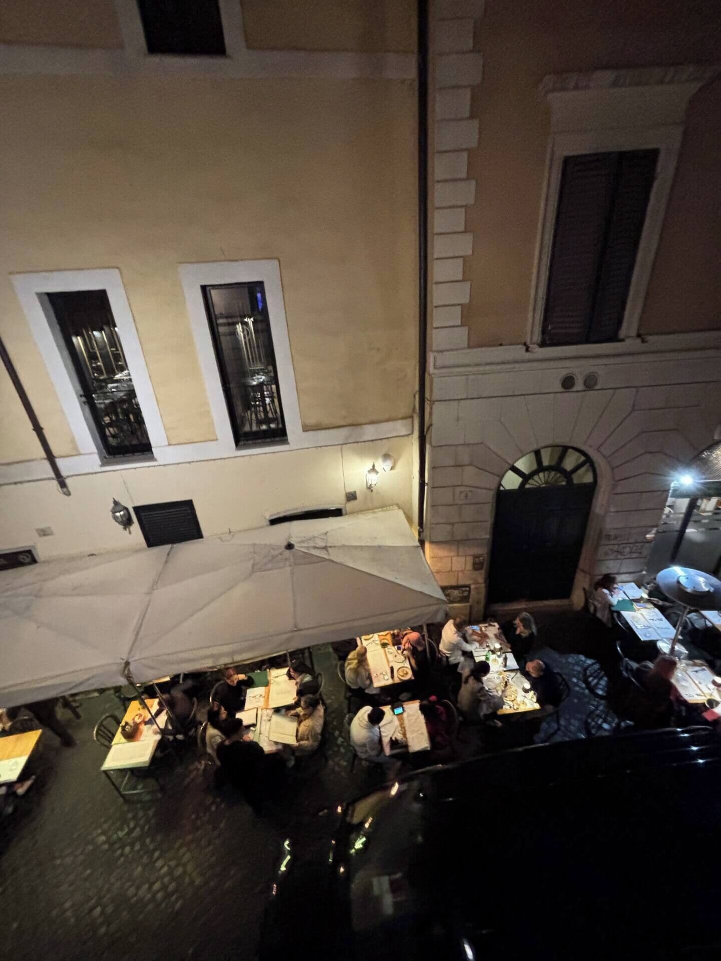 An outdoor dining area with tables and chairs, a white umbrella, and a building with a stone facade and windows.
