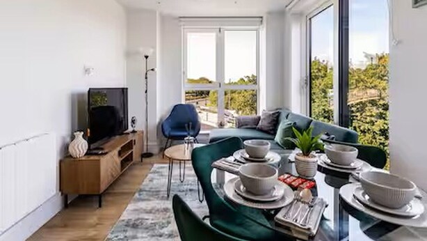 Ein modernes Wohnzimmer mit Fernseher, Sitzgelegenheit, Esstisch und großen Fenstern.