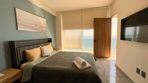A bedroom with a bed, a TV, a large window with a view of the sea, and a door leading outside.