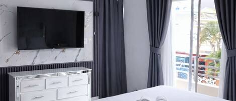 Moderni hotellihuone, jossa on sänky, seinälle asennettu litteänäyttöinen televisio, valkoinen lipasto ja parveke, jossa on verhot.