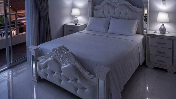 A bedroom with a large bed, two bedside tables, a ceiling-mounted air conditioning unit, and a view through a glass door.