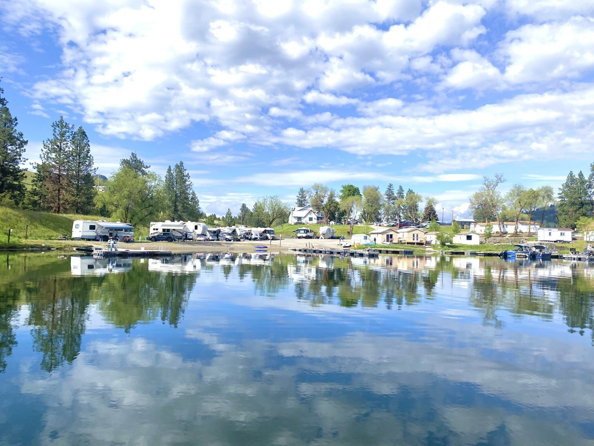 A lakeside RV park with multiple RVs and trailers, calm water reflecting the sky, and surrounding greenery.