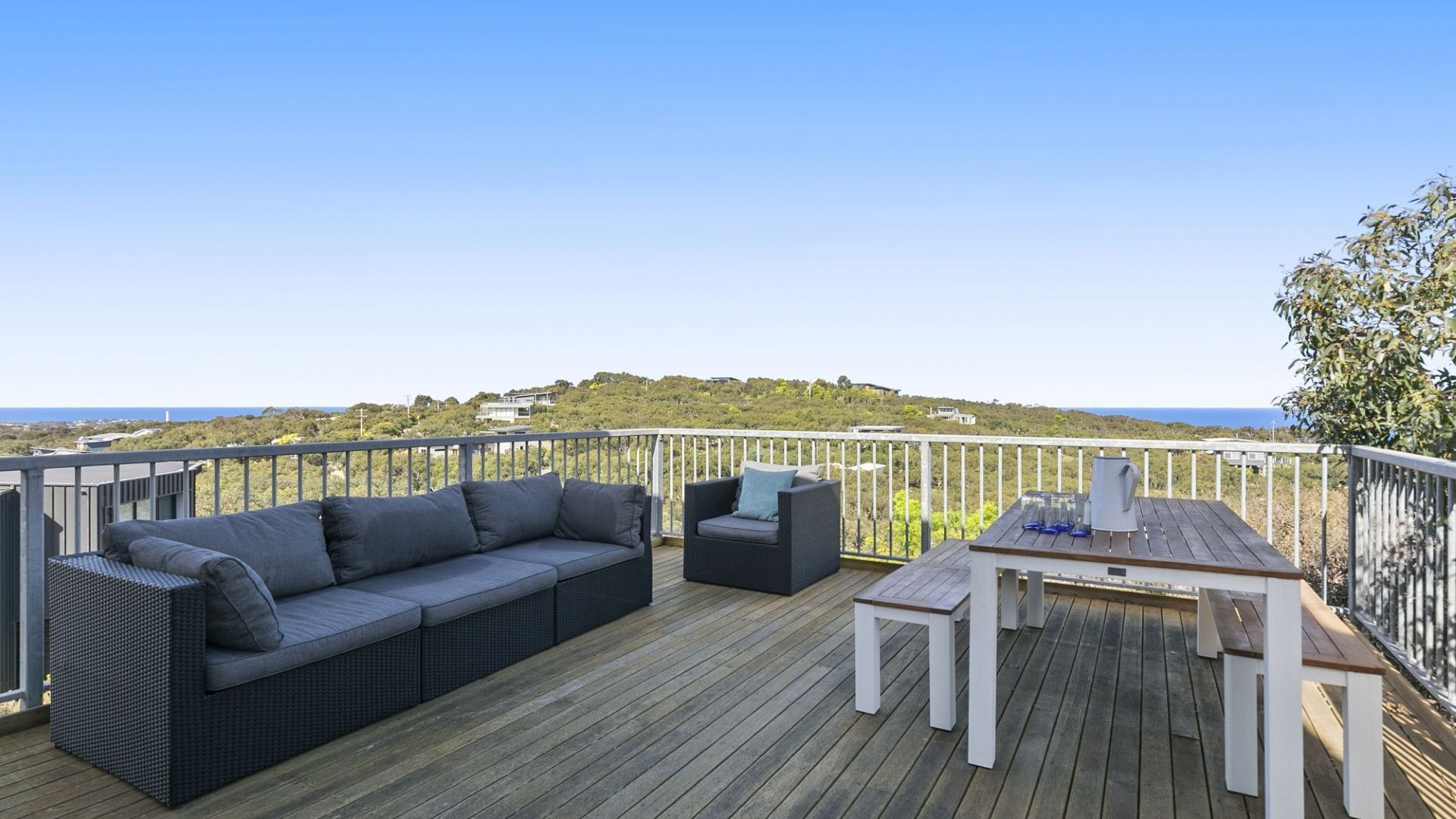 A deck with a sofa, a small table, and a railing overlooking a coastal landscape.