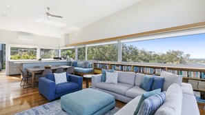 A modern living room with a large window, a built-in bookshelf, and a dining area. - Timbara Hideaway - Ocean Views - Pet Friendly (Fairhaven)