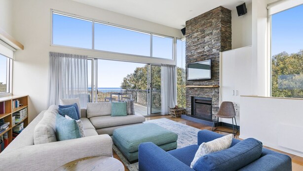 A spacious living room with a large window, a stone fireplace, and a balcony with a view. - Timbara Hideaway - Ocean Views - Pet Friendly (Fairhaven)