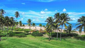 Un complejo turístico en la playa con palmeras, una cabaña de techo de paja y una vista despejada del océano.