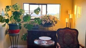 Une pièce avec une fenêtre, une plante en pot, un vase de fleurs, une table recouverte d’une nappe décorative et une lampe.
