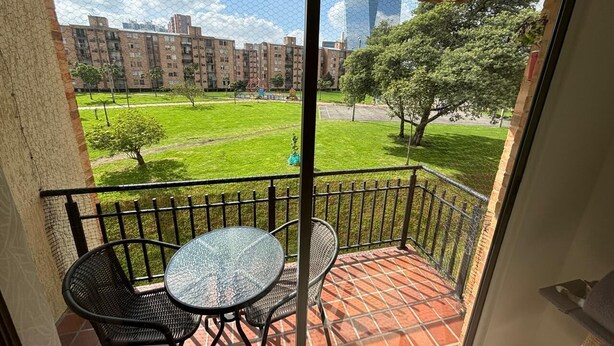 A balcony with a table and chairs, a view of a green park, and a distant building complex. - Cozy, family and pet-friendly apartment, located right beside a beautiful park. (Bogotá)