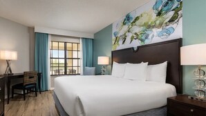 A hotel room with a large bed, two bedside tables, a desk with a chair, a window with curtains, and a wall with a floral painting. - 2 bedroom near Disney with pools and golf courses on the property. (Kissimmee)
