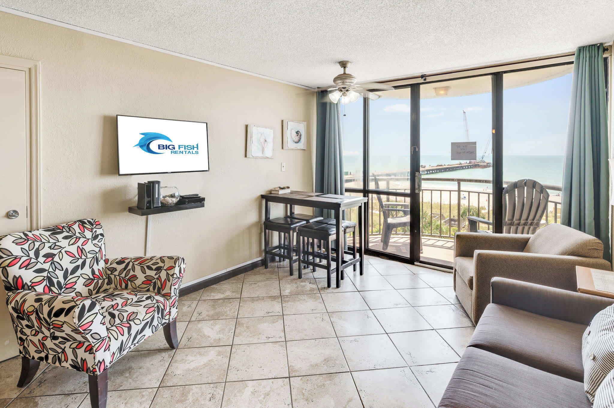 A furnished room with a large window, a ceiling fan, a flat-screen TV, and a view of the ocean.