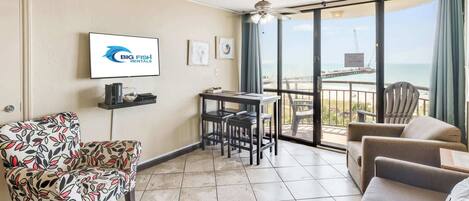 A furnished room with a large window, a ceiling fan, a flat-screen TV, and a view of the ocean.