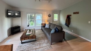 Ein Wohnzimmer mit Sofa, Couchtisch, Fernseher und einem Fenster mit Blick auf Bäume.