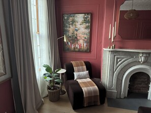 Ein Zimmer mit Kamin, einem Gemälde, einem Sessel mit Decke und einer Topfpflanze.