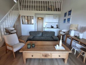 Un salon avec une table basse en bois, un canapé gris, une chaise blanche, un escalier et, au fond, un coin cuisine.