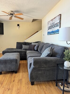 A living room with a sectional sofa, a ceiling fan, a staircase, and a wall-mounted lamp.