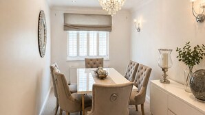 Une salle à manger avec un lustre, une table à manger, des chaises, un radiateur et une fenêtre avec des stores.