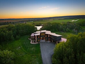 A large, modern house with a spacious driveway, surrounded by lush greenery and a small pond.