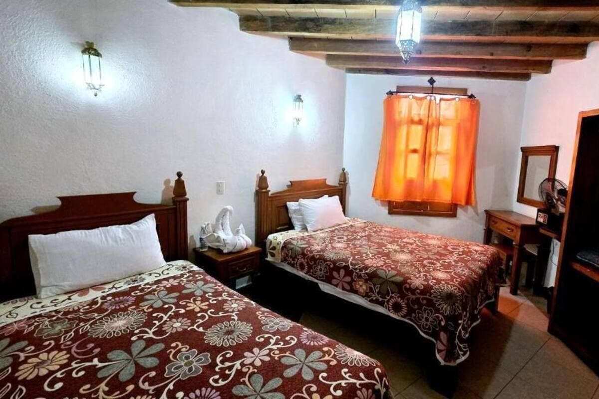 Un dormitorio con dos camas, cabeceros de madera, una mesita de noche, un aparador, un espejo y una ventana con cortinas.