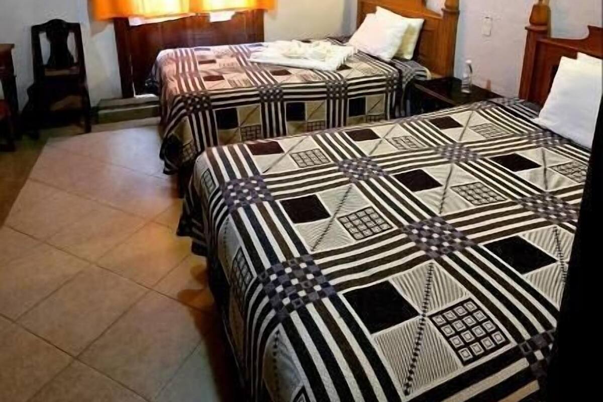 Una habitación con dos camas, vigas de madera en el techo, una ventana con cortinas y una lámpara fijada a la pared.