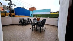 Un porche techado con una mesa redonda y sillas, una planta en maceta y vistas de edificios.