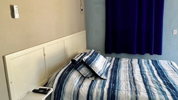A bedroom with a bed, a bedside table, a remote control, a wall-mounted air conditioner, and a curtain. - Cozy Rooms Rocky Point - Walk to Playa Hermosa (Puerto Penasco)