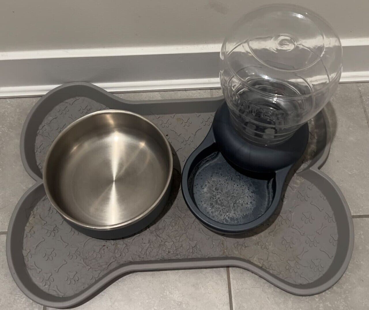 Una estación para alimentar mascotas con un cuenco de metal y un dispensador de agua sobre una bandeja estampada.